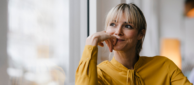 Blond woman in yellow shirt looking out a window pensively